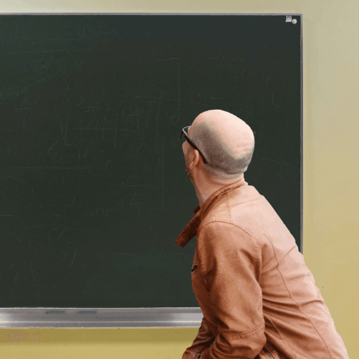 Osoba stojí před školní tabulí v učebně a prohlíží si obsah napsaný křídou. Fotografie znázorňuje profesní rozvoj pedagogických pracovníků, vzdělávání učitelů nebo pedagogické vedení ve školním prostředí.