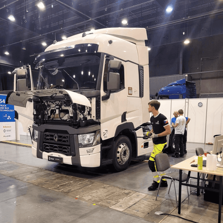 Bílý nákladní tahač Renault s otevřenou přední kapotou stojí v hale na odborné soutěži nebo výstavě. Vedle vozidla se pohybují účastníci a v pozadí jsou další nákladní automobily a pracovní stanoviště.